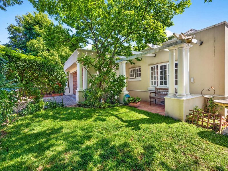 A charming rear garden featuring a paved patio area with a pergola, classical columns, and a well-maintained lawn.