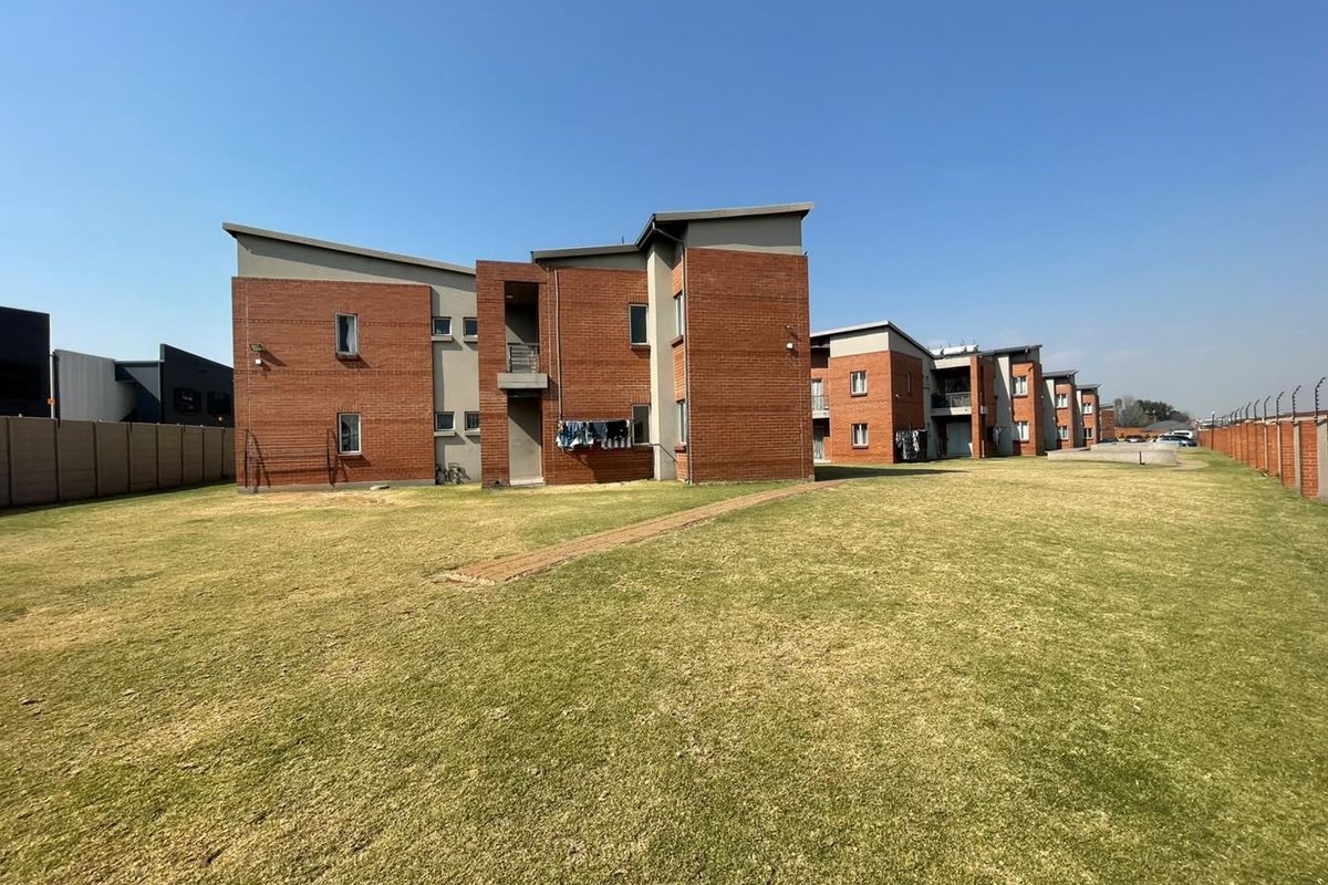A view of the rear elevation of a multi-story apartment complex featuring face brick construction, a large communal lawn, and perimeter security fencing.