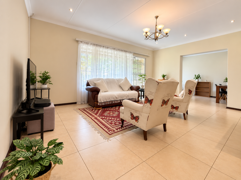 A bright and spacious living room featuring neutral tiled flooring, a classic chandelier, and a sliding door with security gate leading to the exterior.