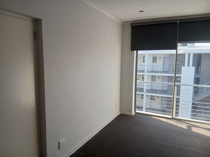 A bright, empty bedroom featuring neutral carpeting, recessed lighting, and a sliding glass door with roller blinds providing access to a balcony.