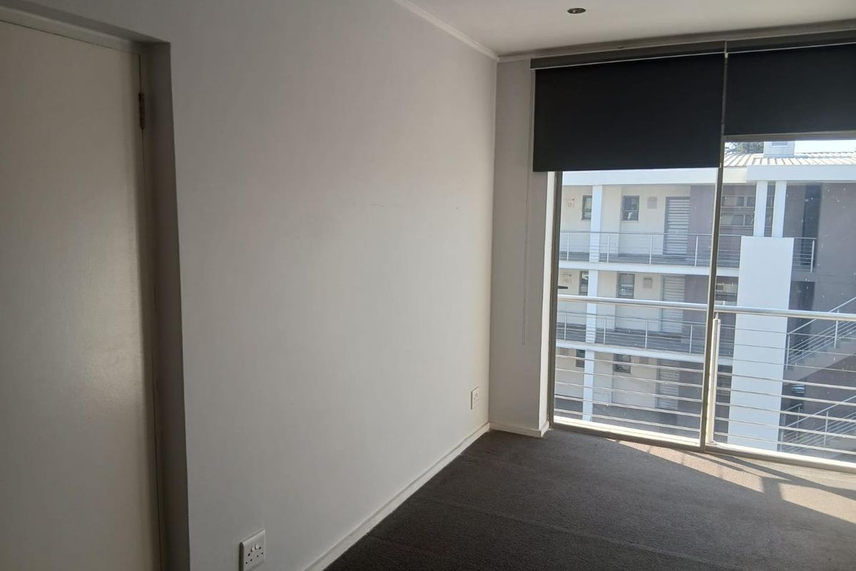 A bright, empty bedroom featuring neutral carpeting, recessed lighting, and a sliding glass door with roller blinds providing access to a balcony.