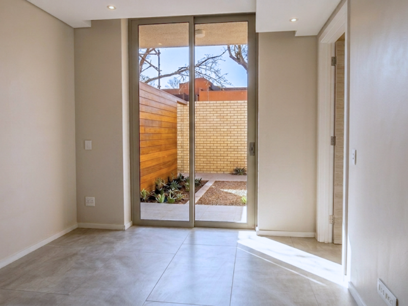 A bright, empty living area featuring large sliding glass doors that open onto a private courtyard with timber cladding and landscaped garden beds.