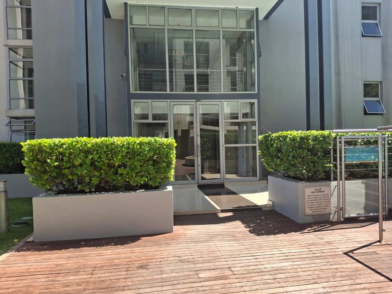 Modern apartment building entrance featuring glass doors, timber decking, and well-maintained raised garden planters.