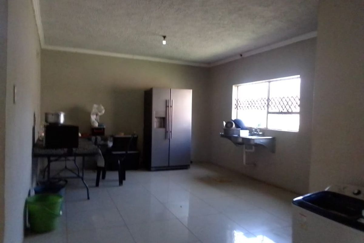 A spacious, sparsely furnished kitchen area featuring tiled flooring, a wall-mounted sink, and large window with security bars.