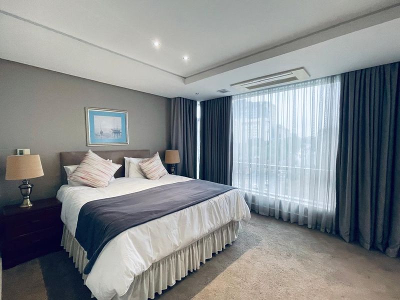 A well-maintained bedroom featuring recessed ceiling lighting, ducted air conditioning, and large floor-to-ceiling windows with elegant drapery.