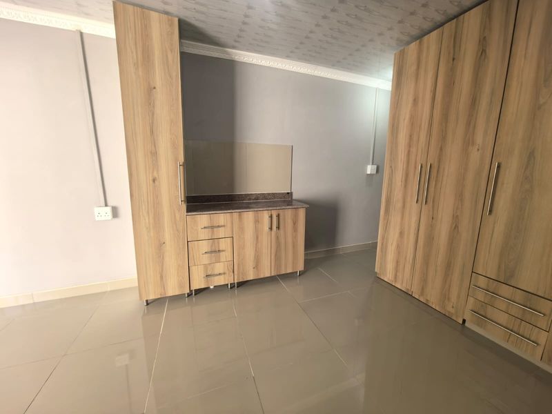 A spacious bedroom featuring modern wood-finish built-in cupboards, tiled flooring, and a neutral color palette, offering a clean and functional living space.