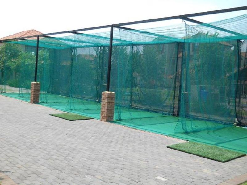 Outdoor cricket practice facility featuring netted bays, brick support pillars, and artificial turf hitting mats on a paved area.