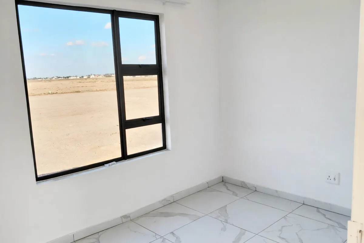 A bright, empty bedroom featuring modern marble-look floor tiles and a large black aluminium window frame with views of the surrounding area.