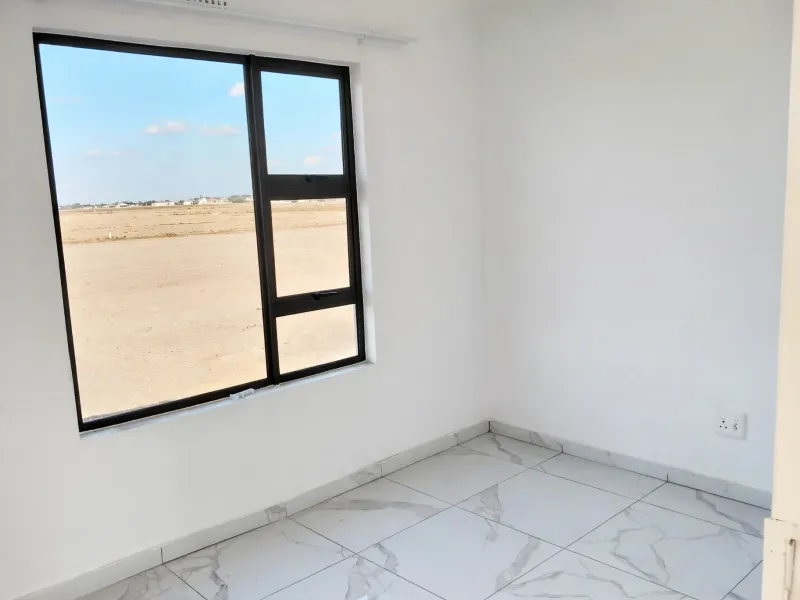 A bright, empty bedroom featuring modern marble-look floor tiles and a large black aluminium window frame with views of the surrounding area.