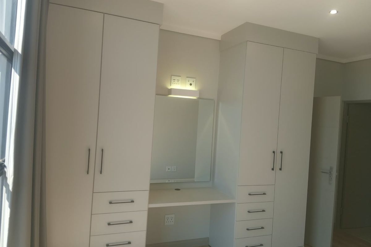 A modern bedroom featuring neutral-toned built-in cupboards with an integrated vanity station, recessed lighting, and light-colored laminate flooring.