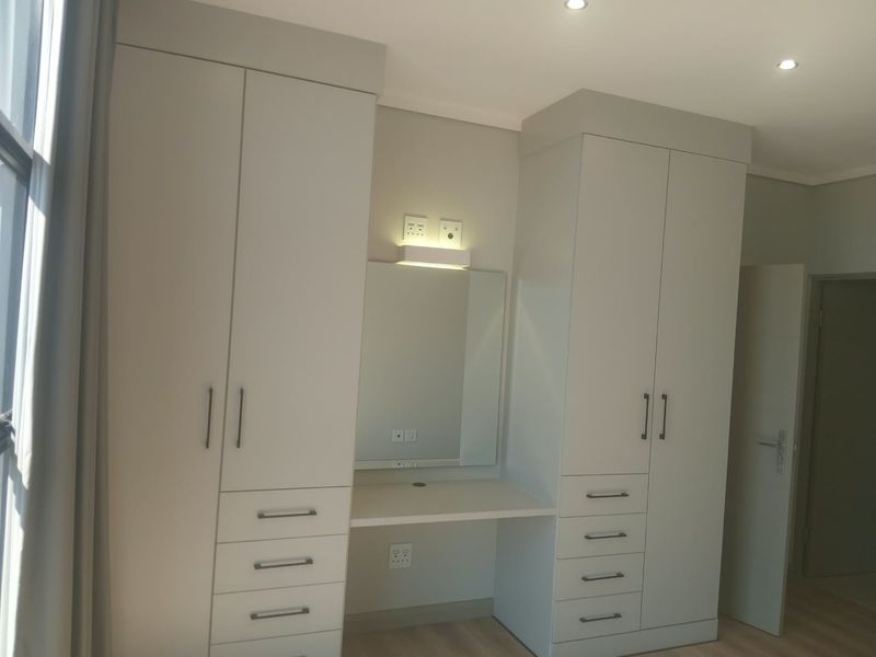 A modern bedroom featuring neutral-toned built-in cupboards with an integrated vanity station, recessed lighting, and light-colored laminate flooring.