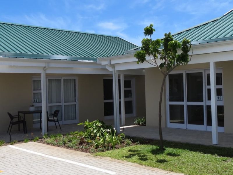 A well-maintained retirement village exterior featuring a paved patio area, green metal roofing, and multiple sliding glass door entrances to individual units.