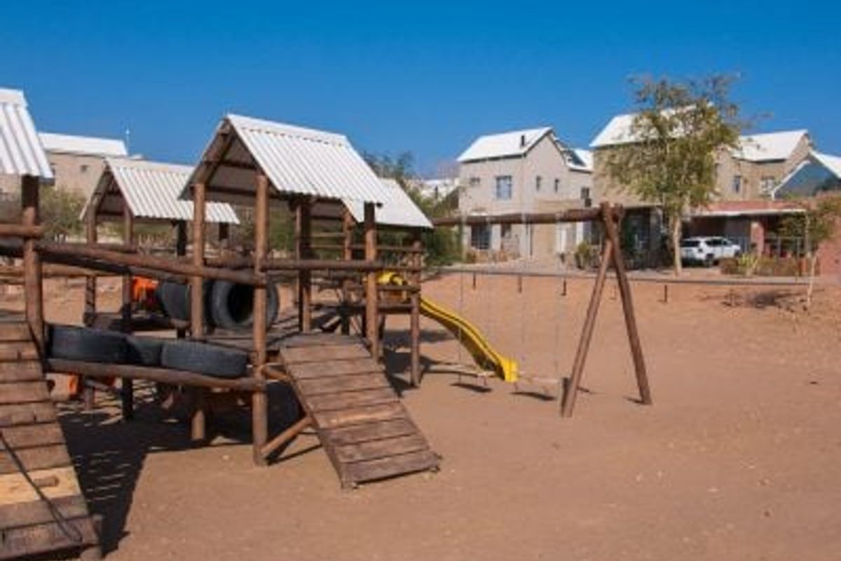 Outdoor communal playground area featuring a wooden climbing structure, swings, and a slide set within a sandy landscape.