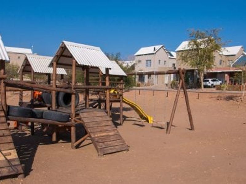 Outdoor communal playground area featuring a wooden climbing structure, swings, and a slide set within a sandy landscape.