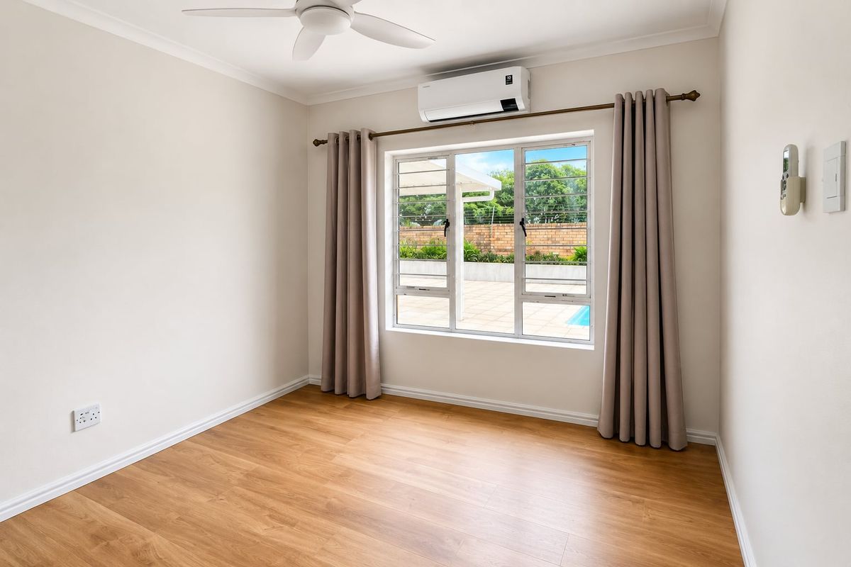 A bright, empty bedroom featuring wood-look laminate flooring, a ceiling fan, and a wall-mounted air conditioning unit overlooking the pool area.