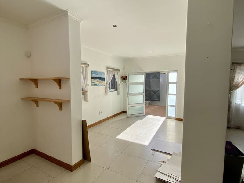 An open-plan living area featuring neutral tiled flooring, recessed lighting, and built-in wall shelving, leading out through a glass-panelled door to an exterior space.