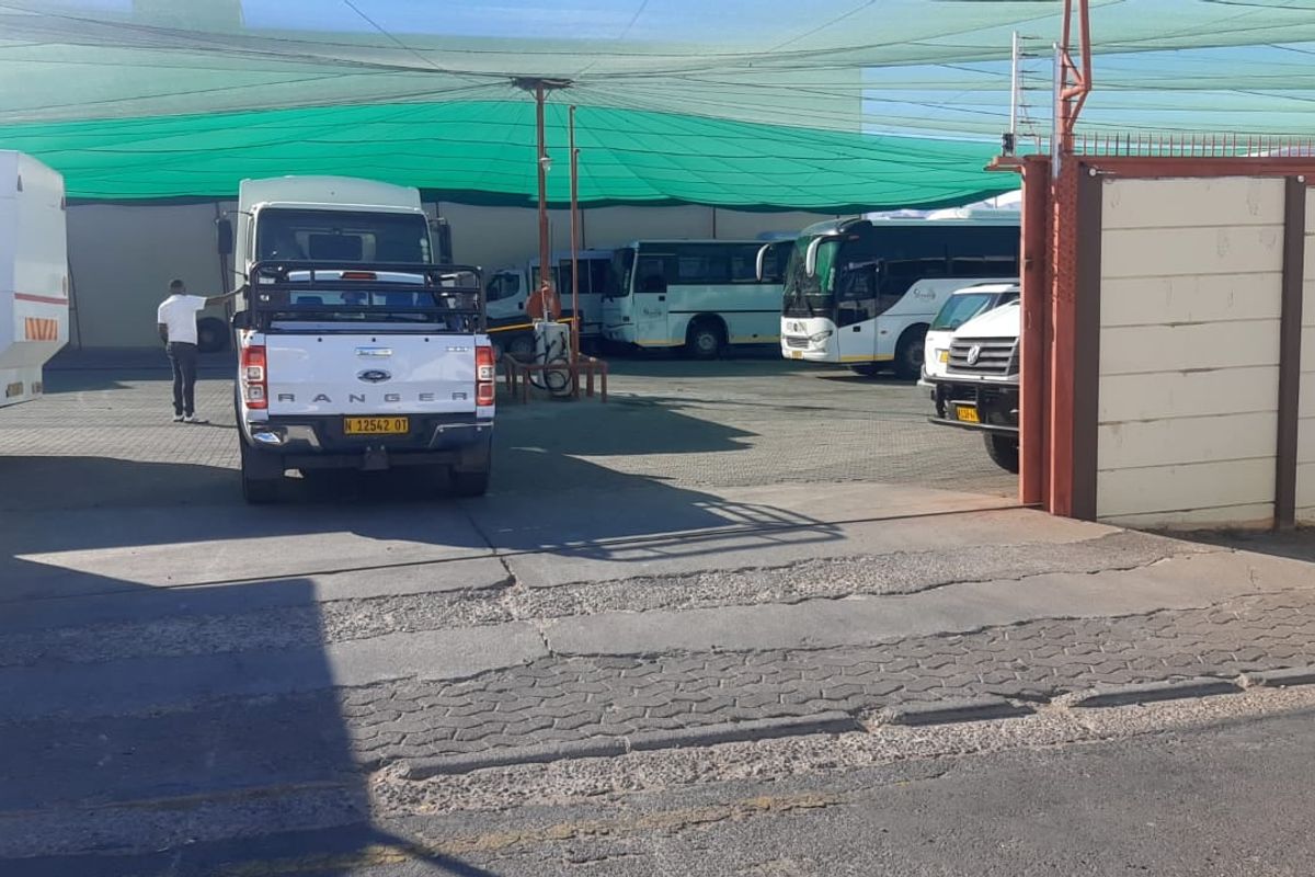 A paved outdoor parking or depot area featuring overhead shade netting, perimeter walling, and a metal gate entrance.