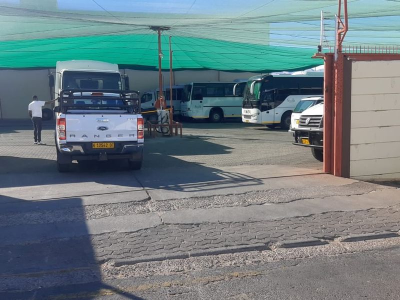A paved outdoor parking or depot area featuring overhead shade netting, perimeter walling, and a metal gate entrance.