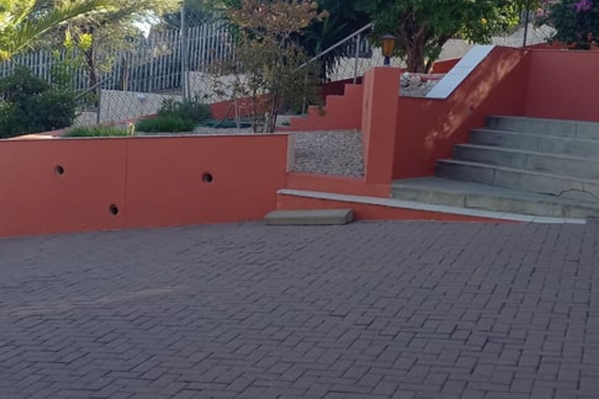 A rear exterior view featuring a paved driveway, tiered retaining walls, and concrete stairs leading up to a landscaped garden area.