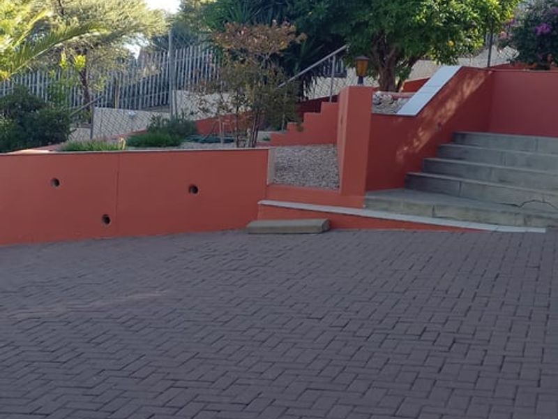 A rear exterior view featuring a paved driveway, tiered retaining walls, and concrete stairs leading up to a landscaped garden area.