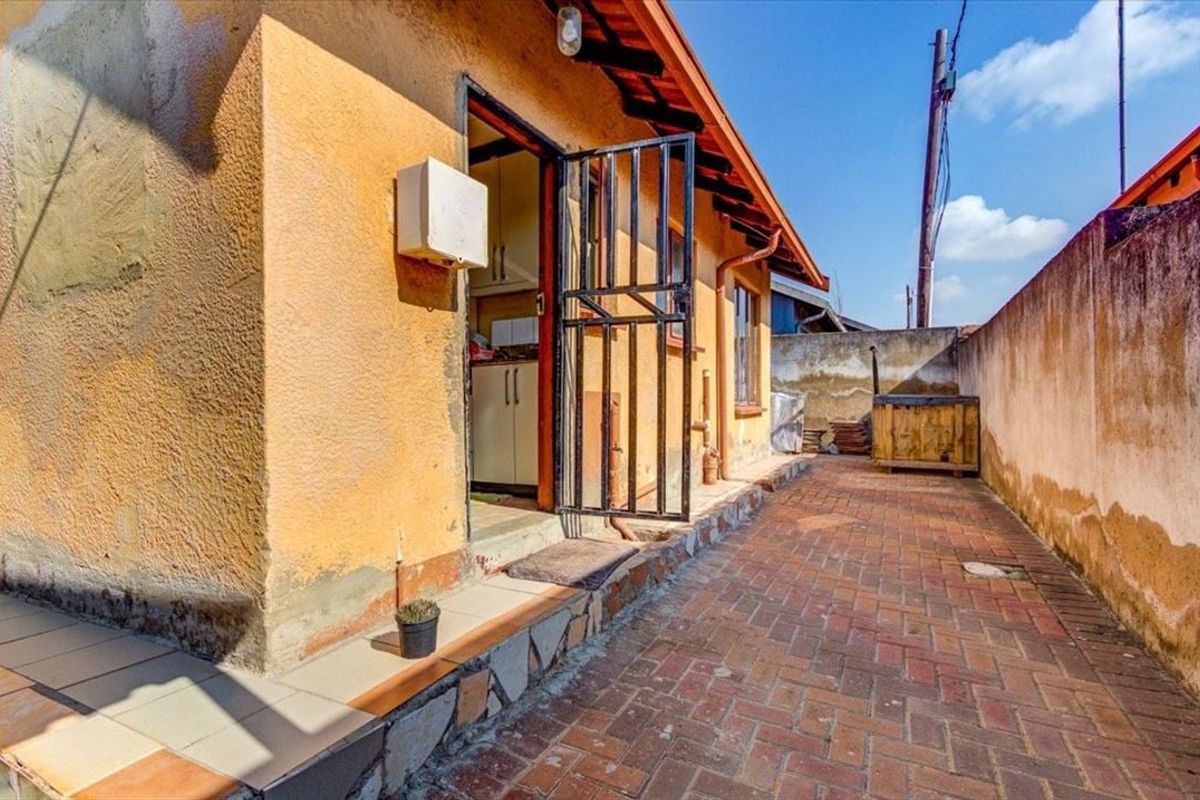 Rear exterior view showing a paved courtyard area with a security gate entrance and weathered wall finishes.
