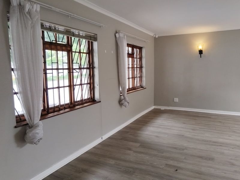 A bright, empty living room featuring modern laminate flooring, neutral wall tones, and timber-framed windows with integrated security bars.