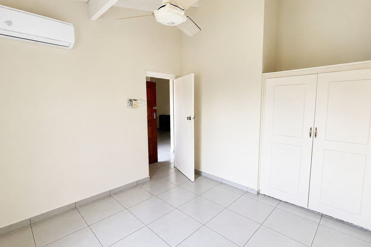 A bright bedroom featuring neutral tiled flooring, a built-in wardrobe, a ceiling fan, and a wall-mounted air conditioning unit.