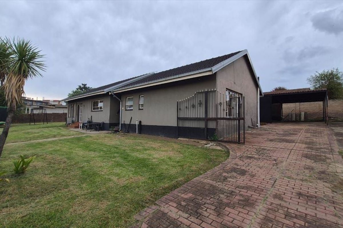 Rear view of the property featuring a paved driveway, carport, and a spacious lawn area with perimeter walling.