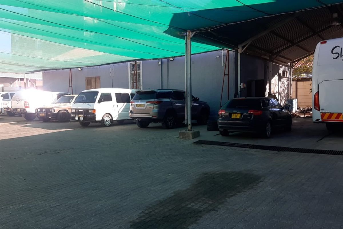A covered outdoor parking area featuring a green shade cloth canopy, paved ground surface, and metal support structures, currently utilized for vehicle storage.