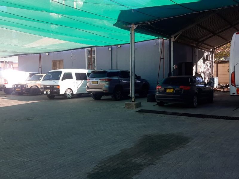 A covered outdoor parking area featuring a green shade cloth canopy, paved ground surface, and metal support structures, currently utilized for vehicle storage.