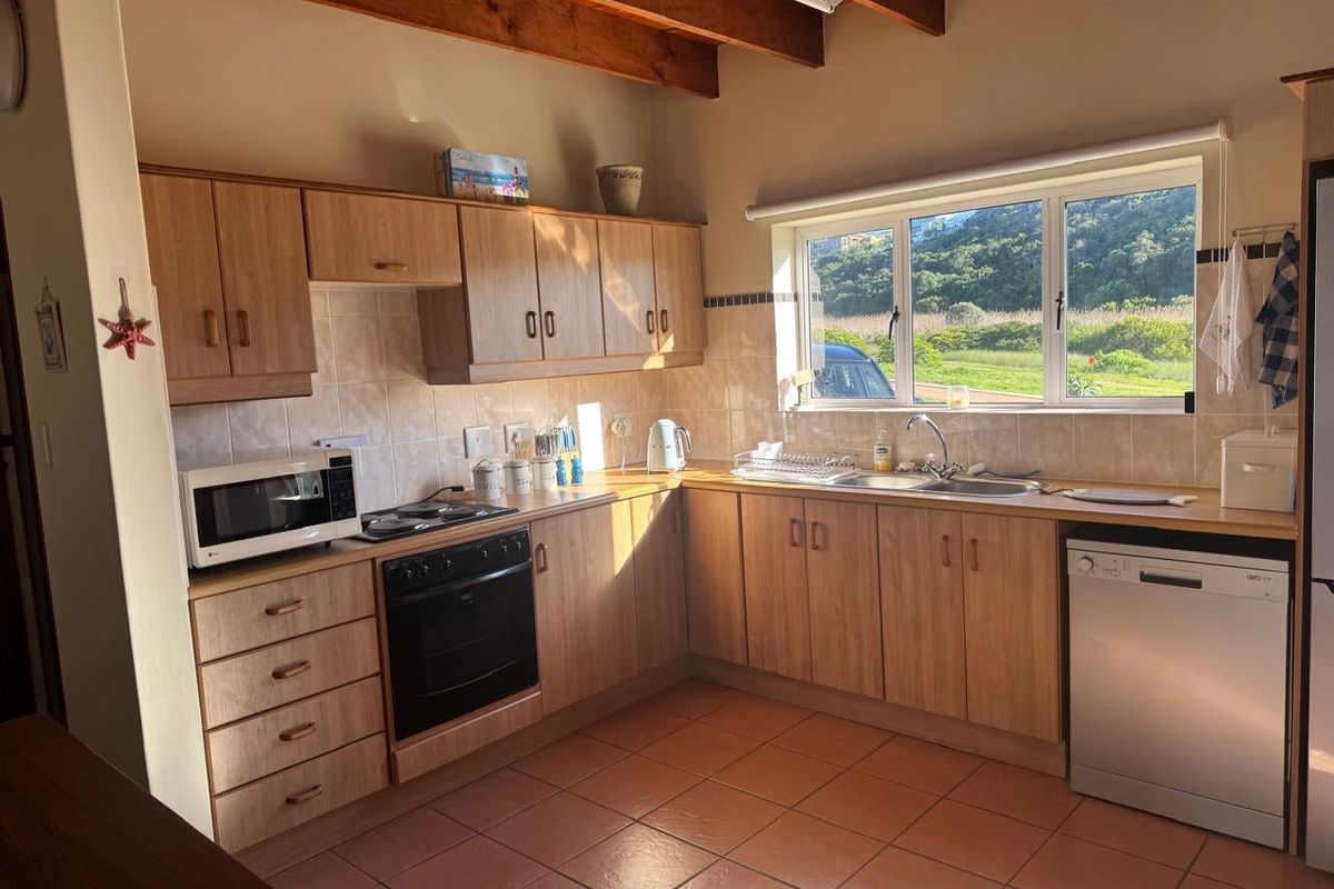 A functional kitchen featuring light wood-look cabinetry, tiled flooring, exposed timber ceiling beams, and a large window providing natural light and garden views.