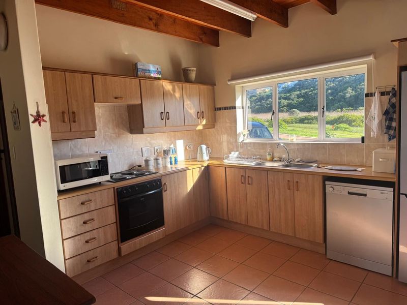 A functional kitchen featuring light wood-look cabinetry, tiled flooring, exposed timber ceiling beams, and a large window providing natural light and garden views.
