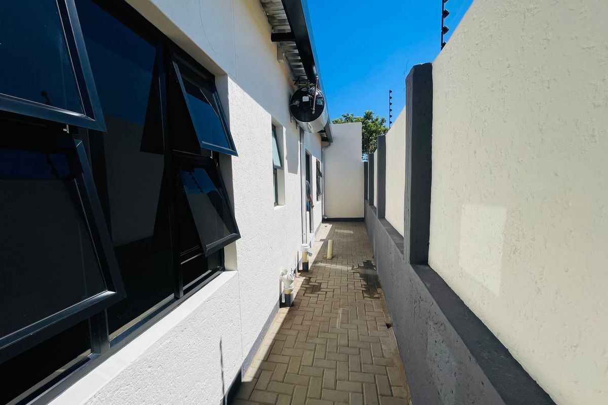 A clean, paved side passage featuring modern aluminium windows, boundary wall with electric fencing, and an external geyser unit.
