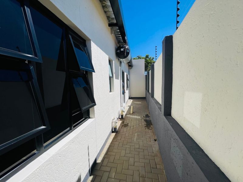 A clean, paved side passage featuring modern aluminium windows, boundary wall with electric fencing, and an external geyser unit.