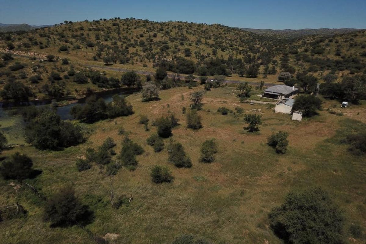 Aerial view of a rural farm property featuring open grazing land, a river, and existing farm structures set against a hilly landscape.