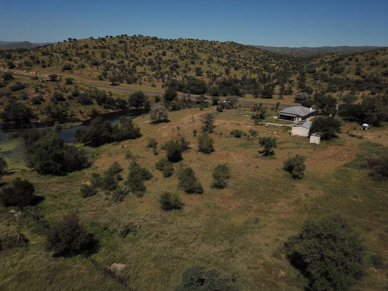 Aerial view of a rural farm property featuring open grazing land, a river, and existing farm structures set against a hilly landscape.