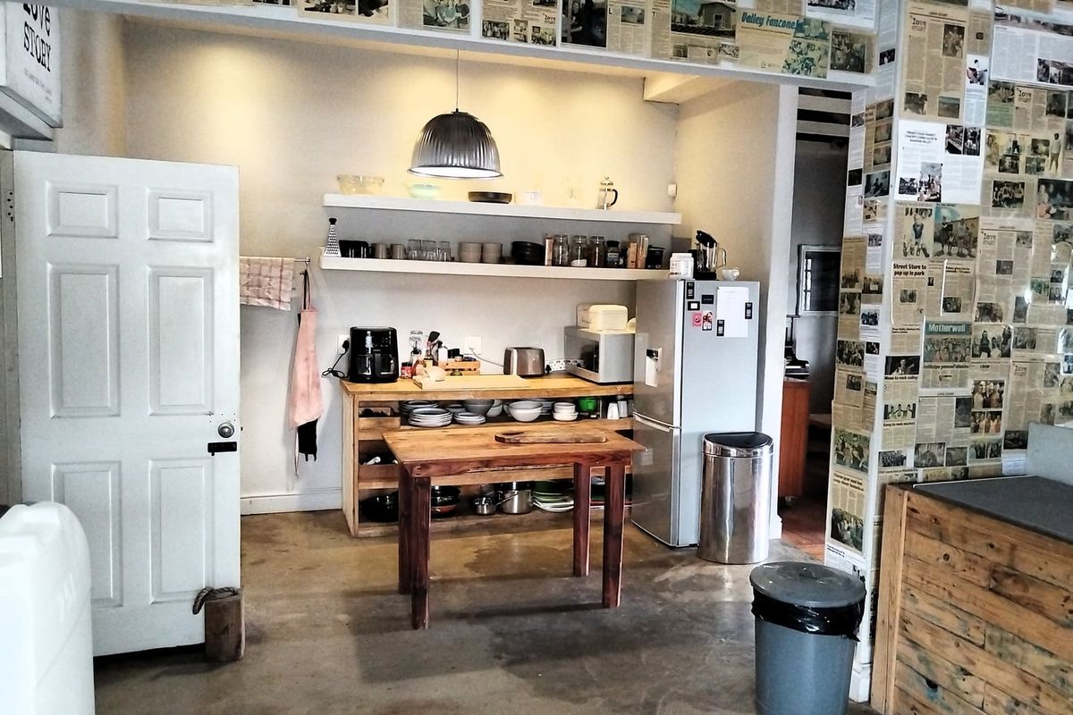 A rustic kitchen area featuring open shelving, concrete flooring, and a central wooden prep table, currently styled with a unique newspaper-covered wall finish.