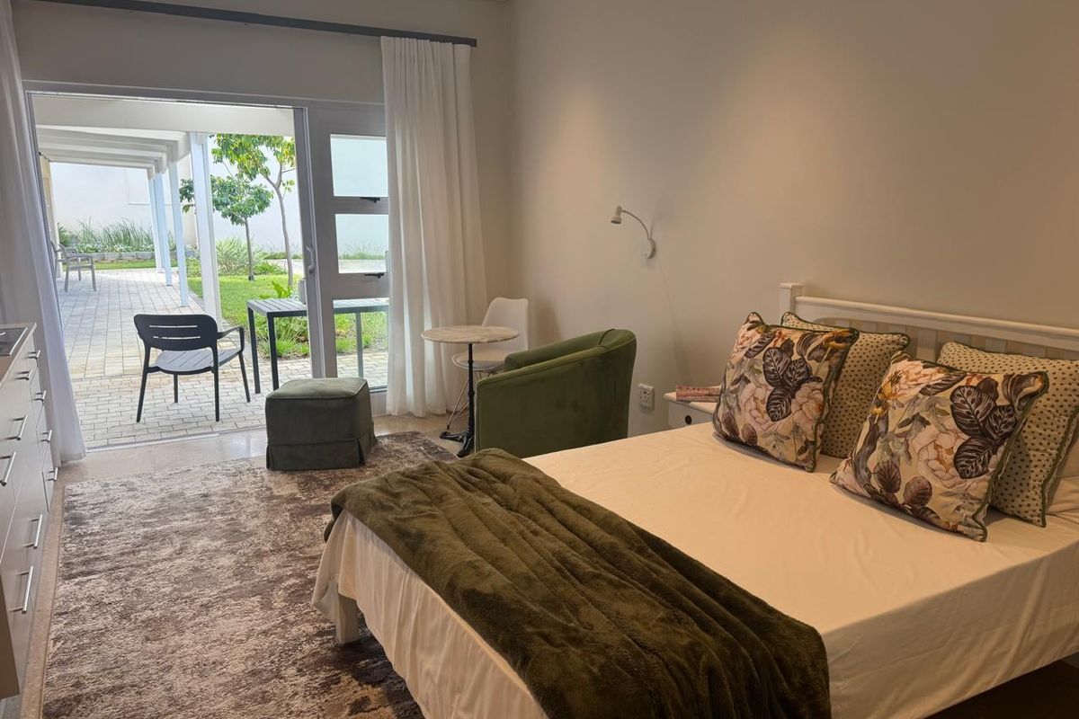 A well-lit bedroom featuring neutral tones, tiled flooring, and a large sliding glass door providing direct access to a paved patio and garden area.