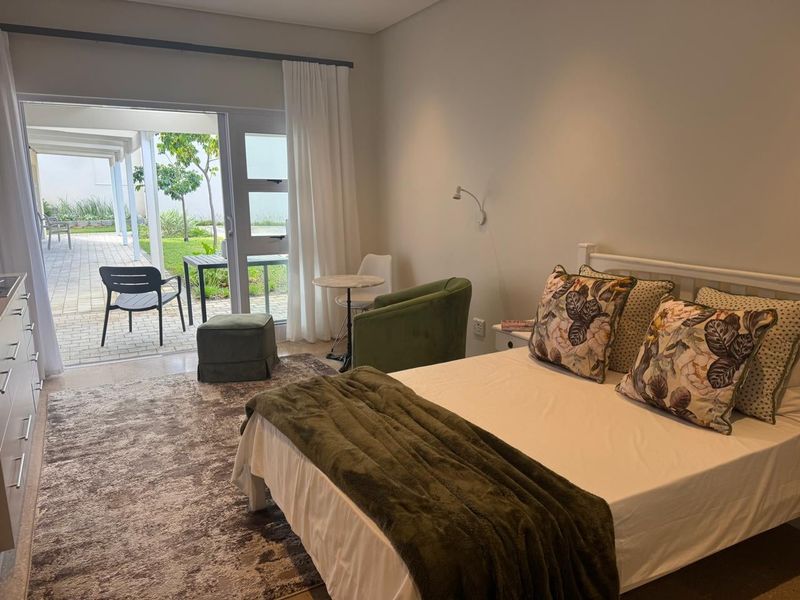 A well-lit bedroom featuring neutral tones, tiled flooring, and a large sliding glass door providing direct access to a paved patio and garden area.