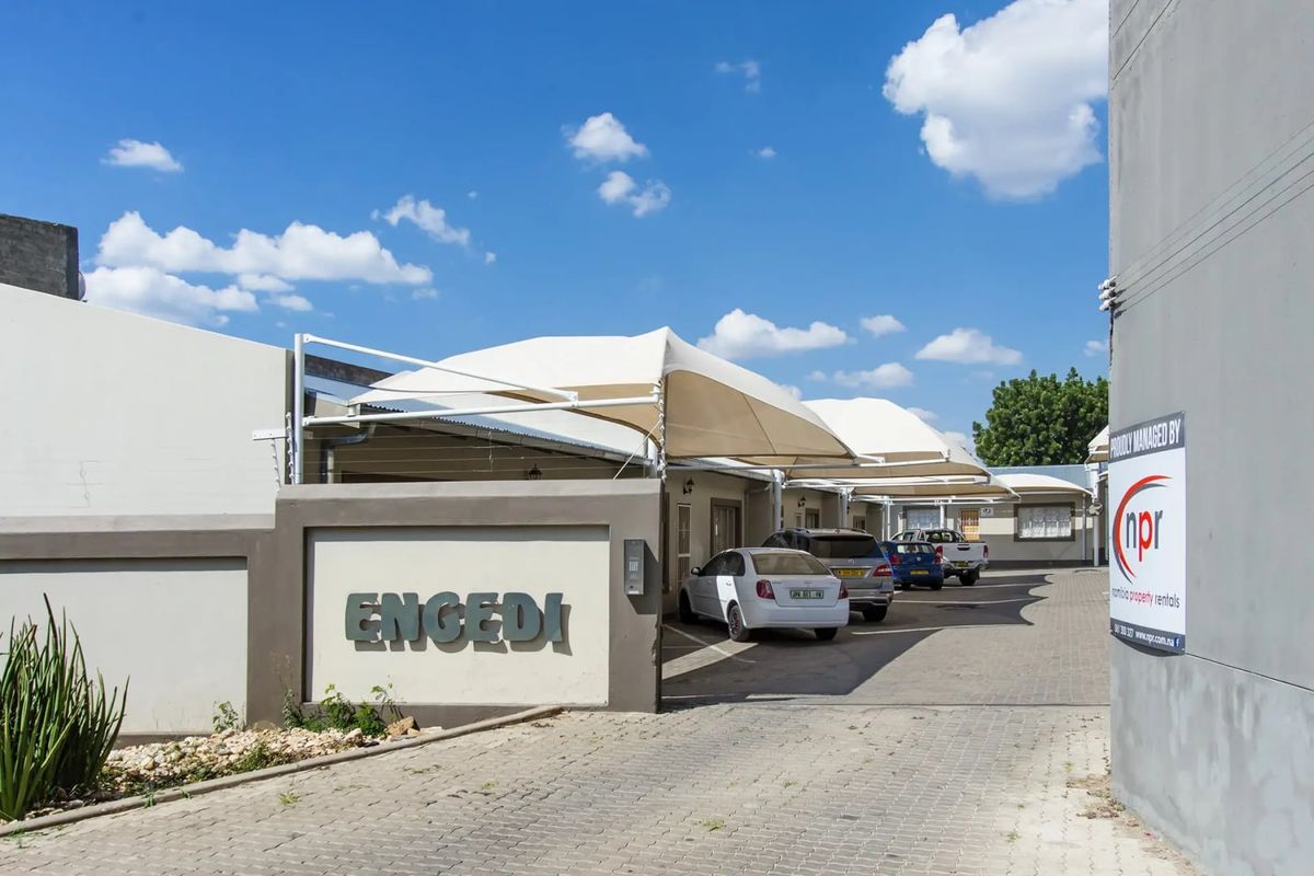 Front entrance view of the townhouse complex featuring a paved driveway, dedicated shade sail parking bays, and clear property signage.