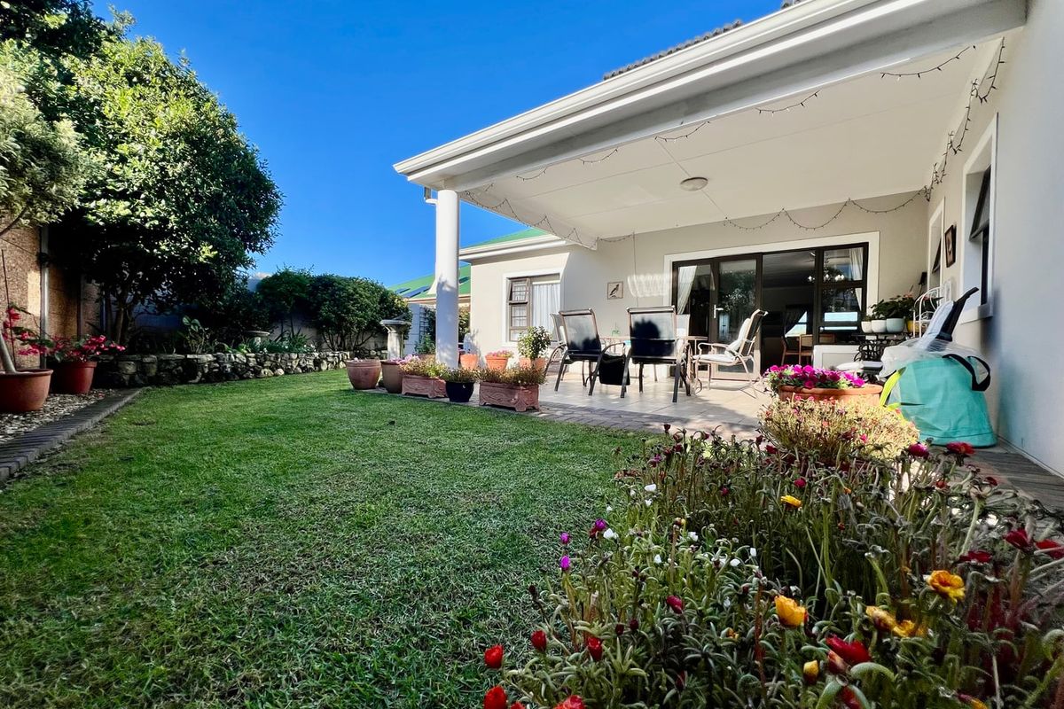 A well-maintained rear garden featuring a covered patio area with paved flooring, accessible via sliding glass doors, and a stone retaining wall.