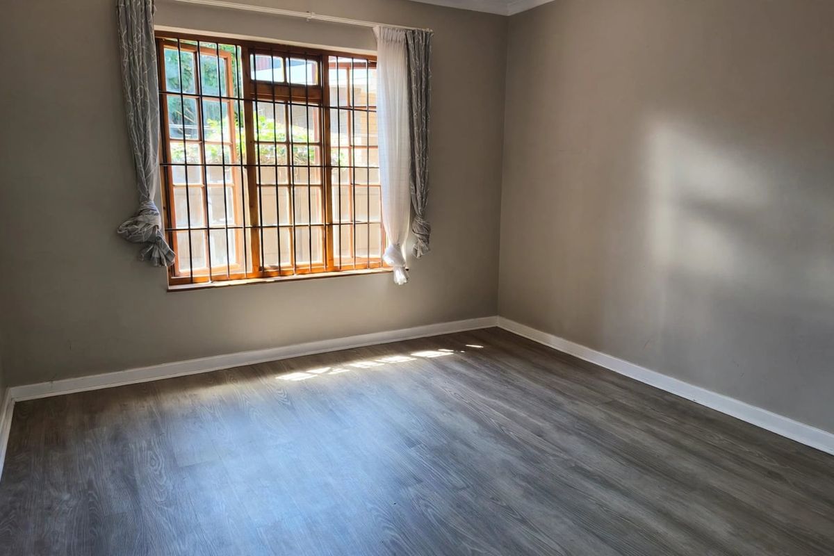 A bright, empty bedroom featuring modern laminate flooring, neutral wall paint, and a wooden window frame fitted with security bars.