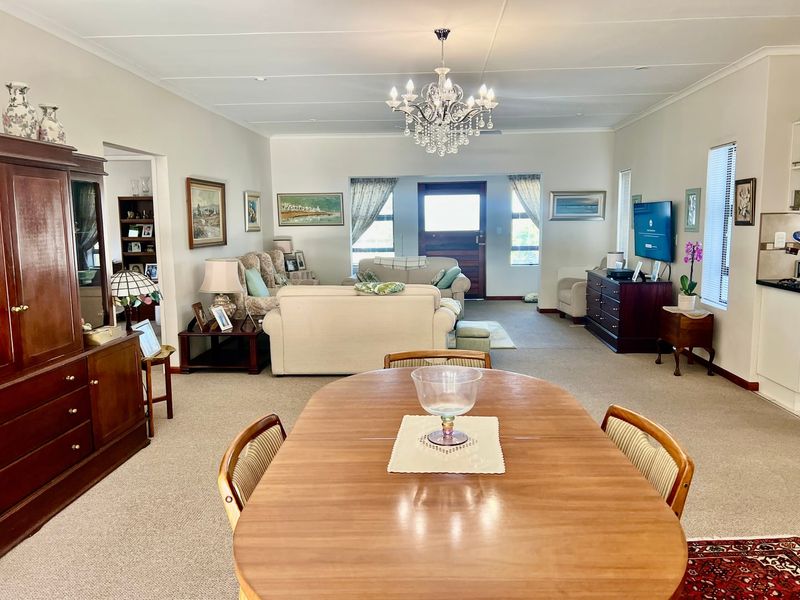 Spacious open-plan living and dining area featuring neutral carpeting, a decorative crystal chandelier, and natural light through front entrance and window openings.