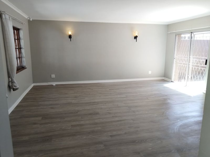 A spacious, empty living room featuring modern laminate flooring, neutral wall tones, wall-mounted lighting, and a sliding glass door leading to an outdoor area.