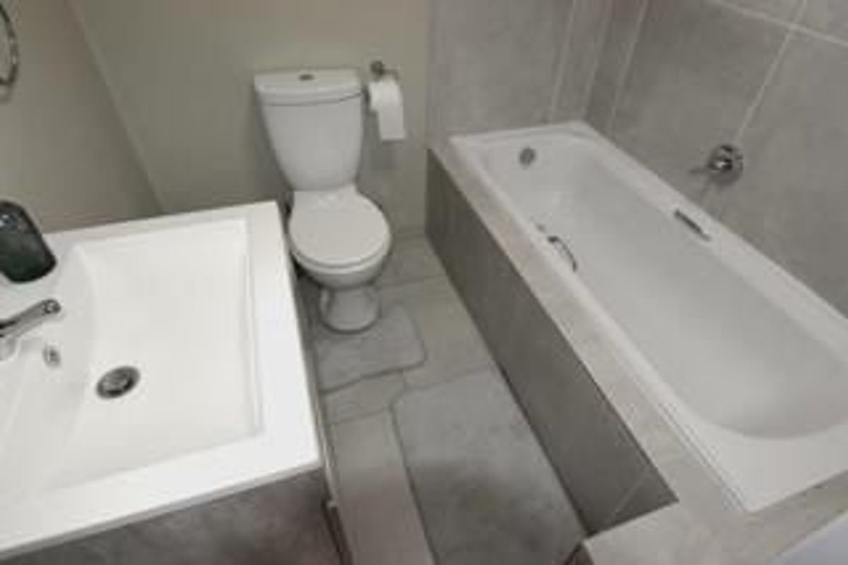 A clean and modern bathroom featuring a built-in bathtub, a white ceramic toilet, and a vanity basin with neutral grey wall and floor tiling.