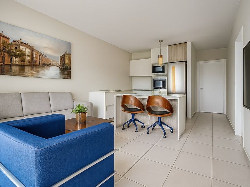 Modern open-plan living area featuring a compact kitchen with built-in appliances, a central island, and neutral tiled flooring throughout.