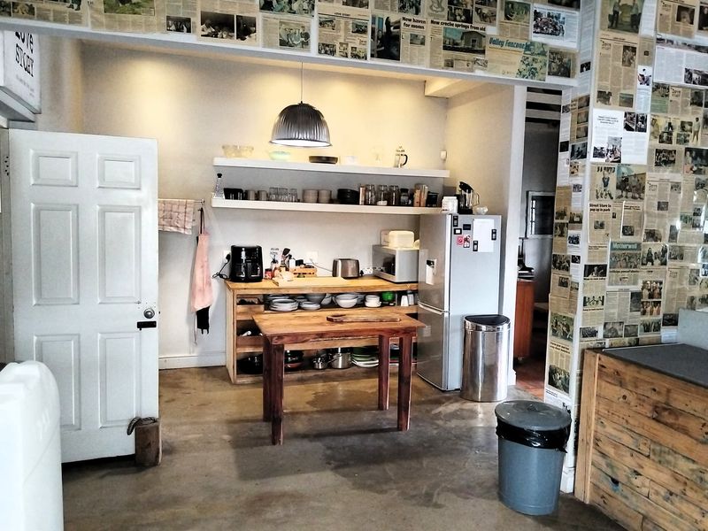 A rustic kitchen area featuring open shelving, concrete flooring, and a central wooden prep table, currently styled with a unique newspaper-covered wall finish.