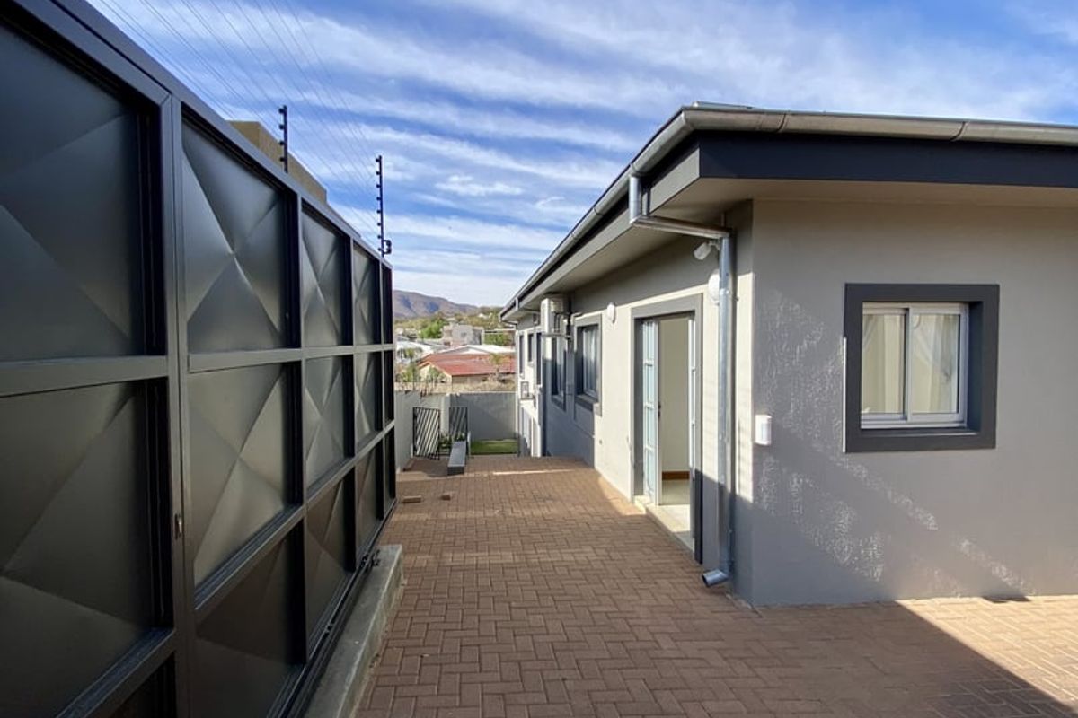A well-maintained rear exterior area featuring paved patio space, modern grey wall finishes, and secure metal boundary fencing with electric security wires.