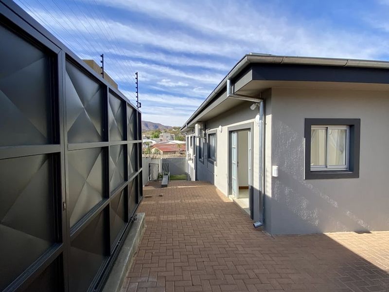 A well-maintained rear exterior area featuring paved patio space, modern grey wall finishes, and secure metal boundary fencing with electric security wires.