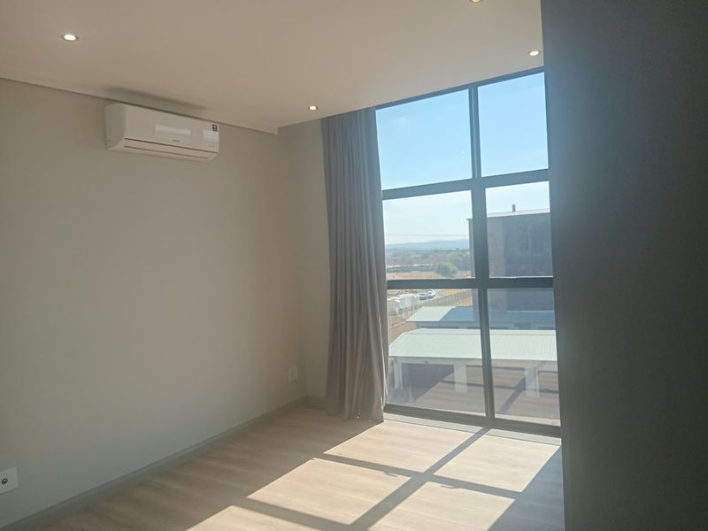 A bright, modern, and empty bedroom featuring laminate flooring, recessed downlights, a wall-mounted air conditioning unit, and a large window with natural light.